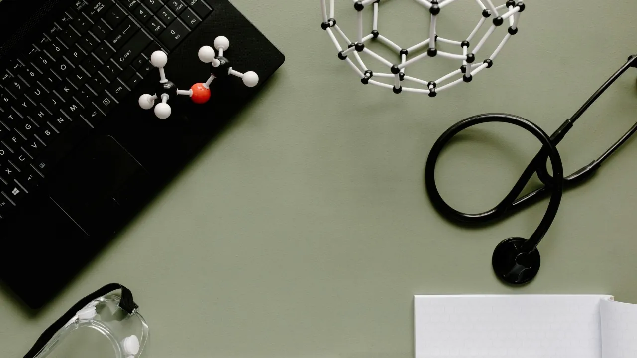 A laptop, stethoscope, molecular model and safety glasses on a green desk.