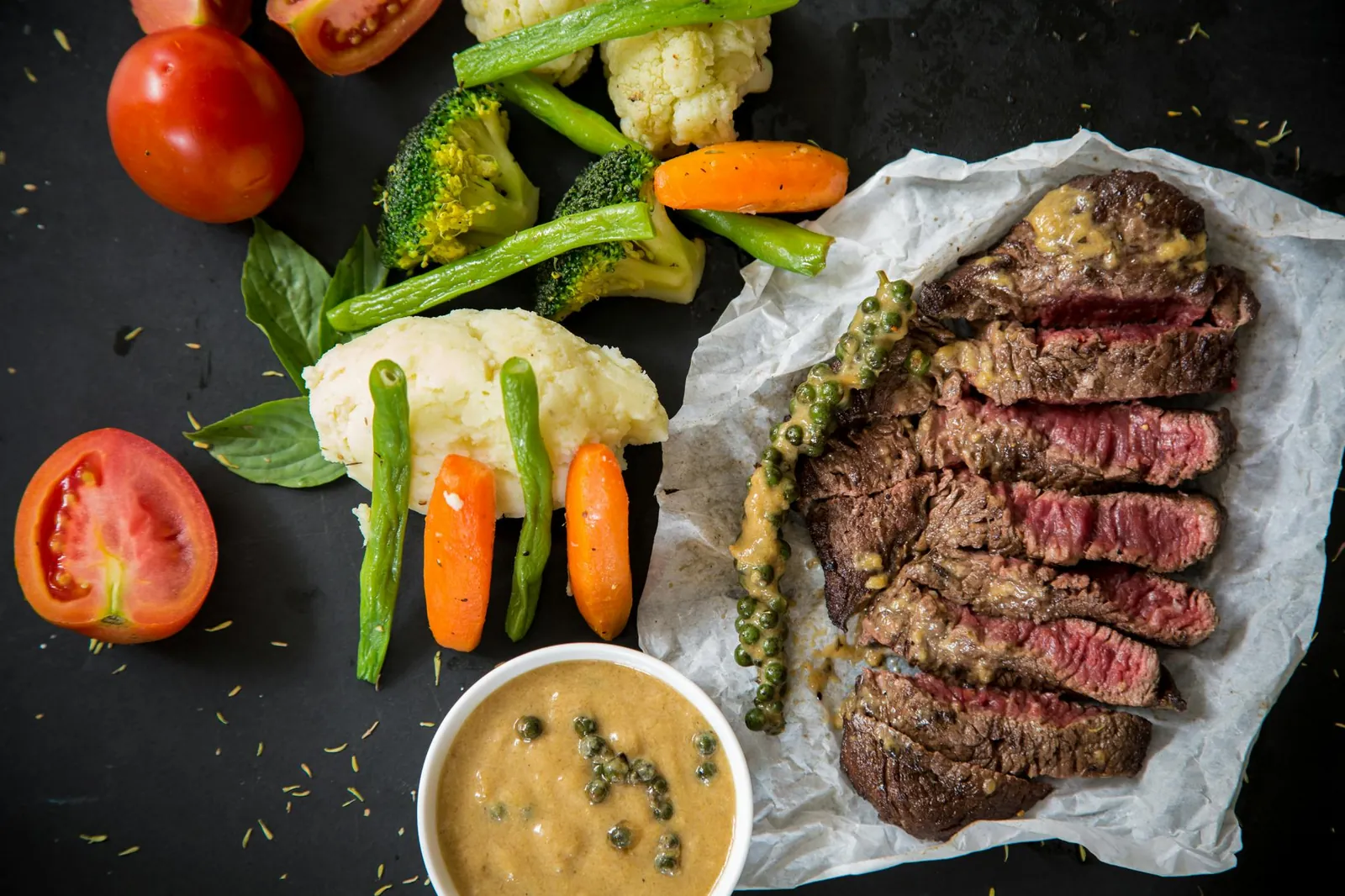 Sliced grilled steak beside roasted vegetables, tomatoes and a bowl of sauce on a dark table.