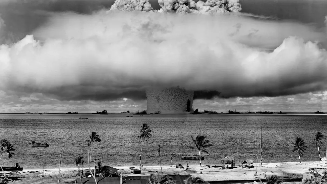 Black-and-white photo of a nuclear test cloud rising over the sea, seen from a palm-lined shore.